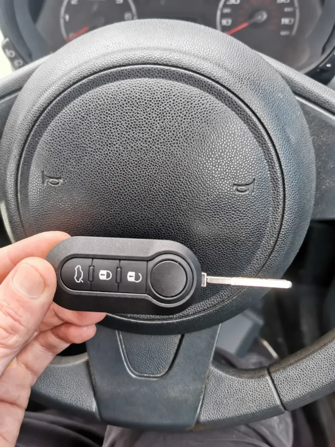 Hand holding a three-button flip key with its blade extended beside a steering wheel